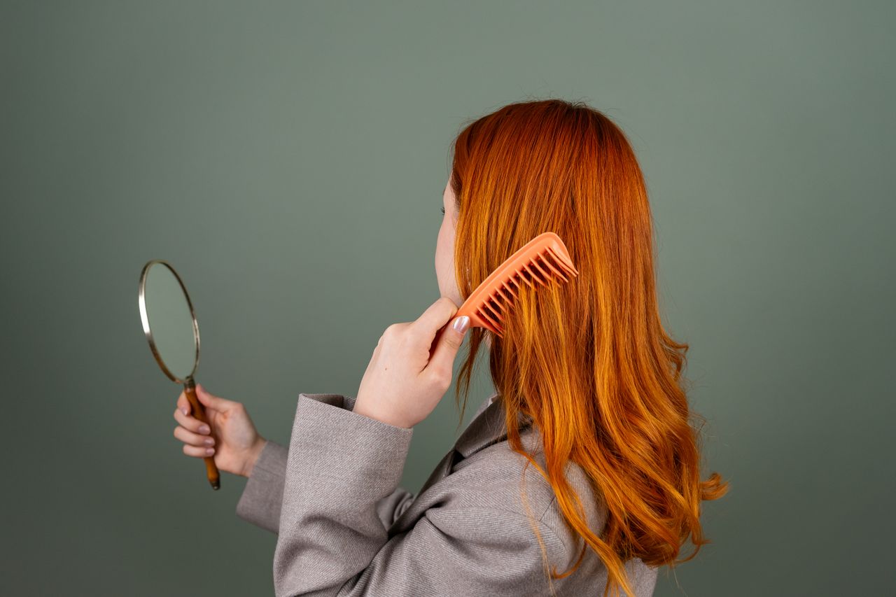 services-04 Red-haired woman using a comb while holding a mirror, focusing on hair care and beauty.